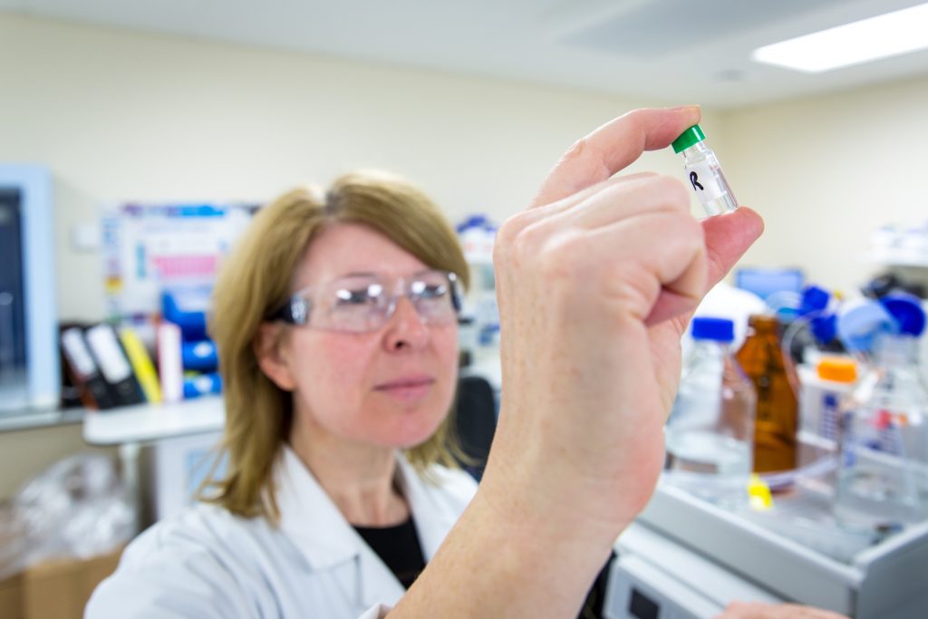 Image shows a female scientist holding up a vile of liquid in a lab.