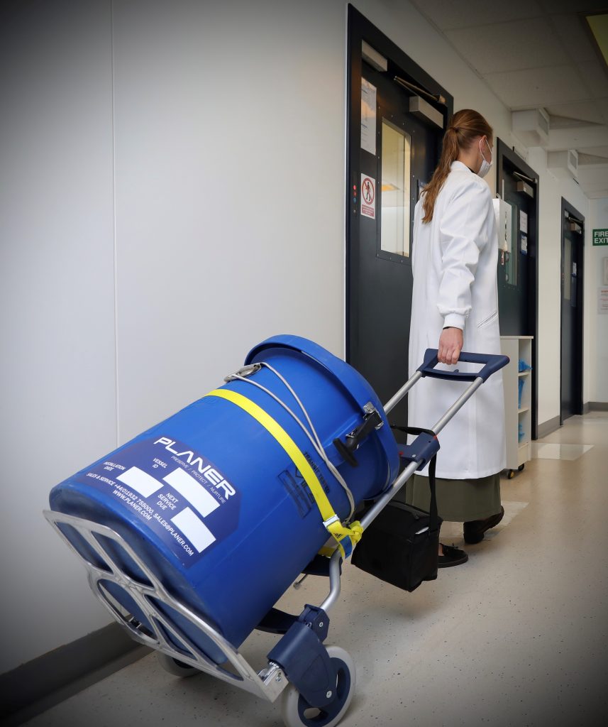 The image shows a female scientist in a white lab coat and protective mask walking along a corridor pulling a blue cylinder container on a trolley.  