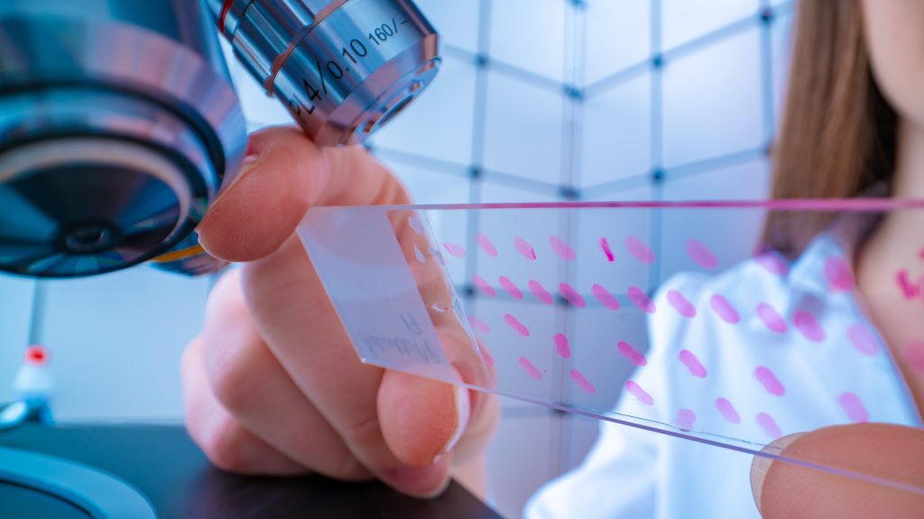 The image shows a microscope with a slide underneath it, held by a female scientist in a lab coat.