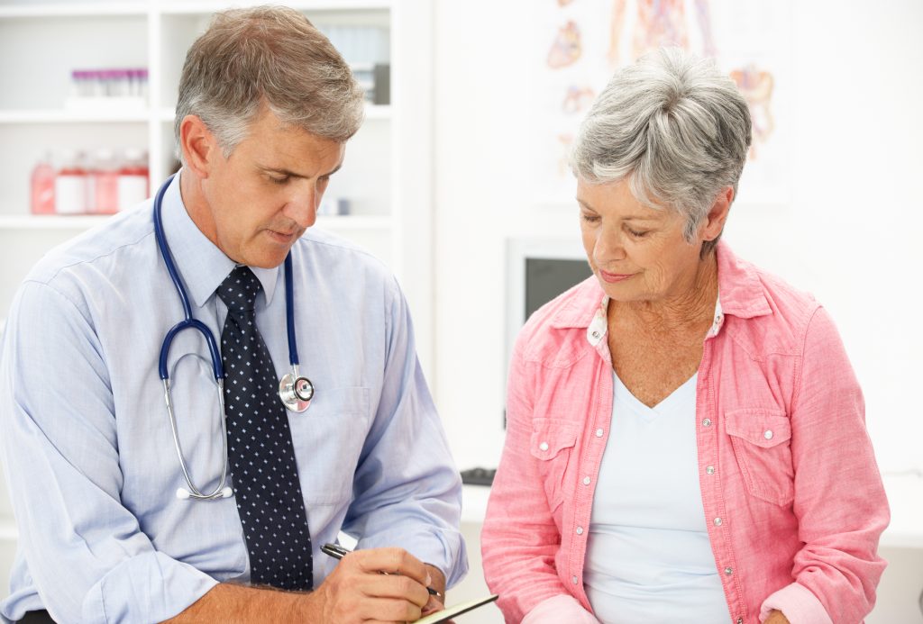 The image shows a male GP sitting with a female patient, making notes.