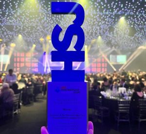 A bright blue award saying HSJ is held up with a back drop of a glittering awards ceremony.