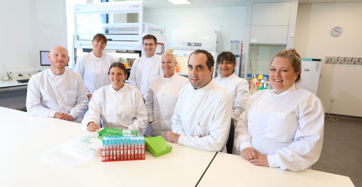 A picture of the lab team sitting together in the lab smiling, with a collection of scientific items placed on the table in front of them.