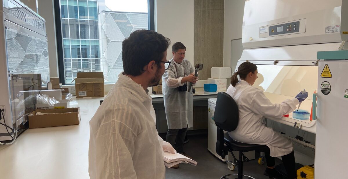 Journalist watching the lab team at work, wearing protective clothing. There is a man holding a camera filming a scientist working.