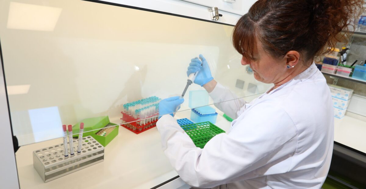 A scientist with a device in a test tube, with lots of racks for test tubes in front of her, some already full.