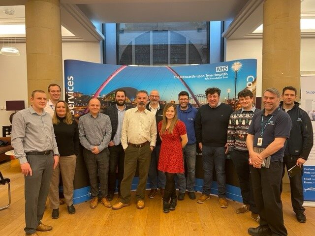 The image shows Charlotte Fox who leads the innovation team with guests in front of a banner stand at an innovation event.