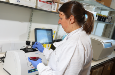 The image shows a female scientist holding a dish containing a liquid and putting a drop onto a piece of equipment.