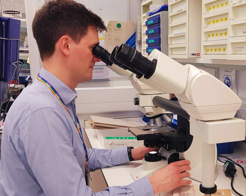 A man looking down a microscope, wearing a blue striped shirt and an NHS lanyard.