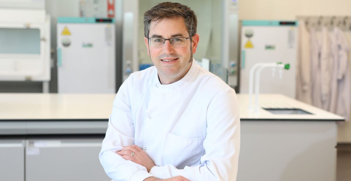 Portrait of Jamie Burbeck sat at a bench in a lab, wearing a white lab coat.