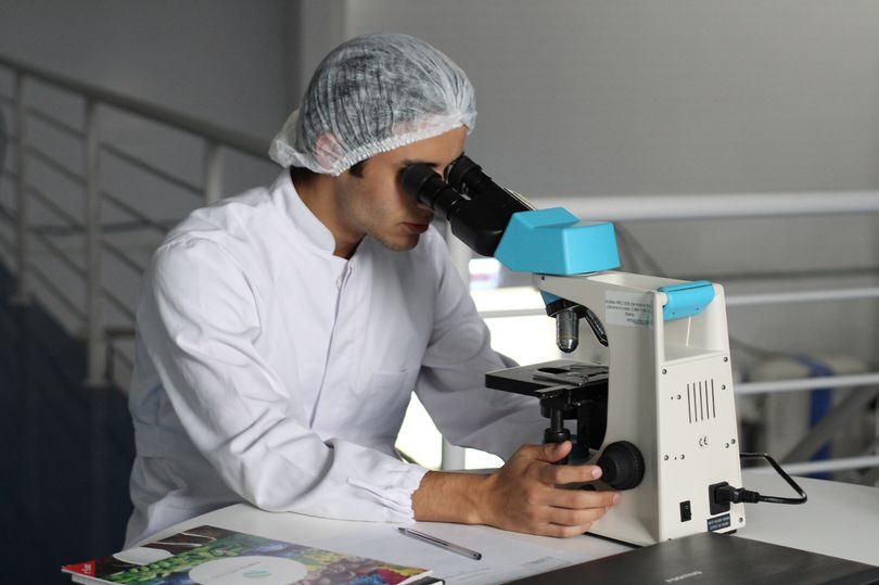 A scientist wearing a white lab coat and a protective hat to cover hair whilst looking through a microscope.
