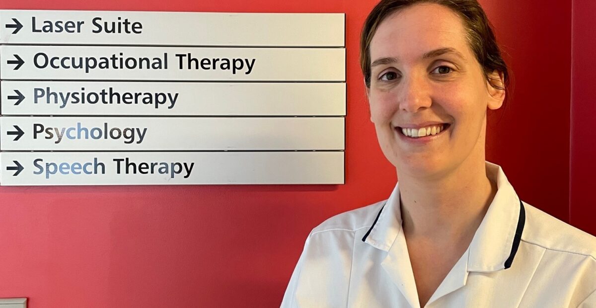 The image shows Suzanne standing in her uniform by a hospital sign for the physiotherapy department.