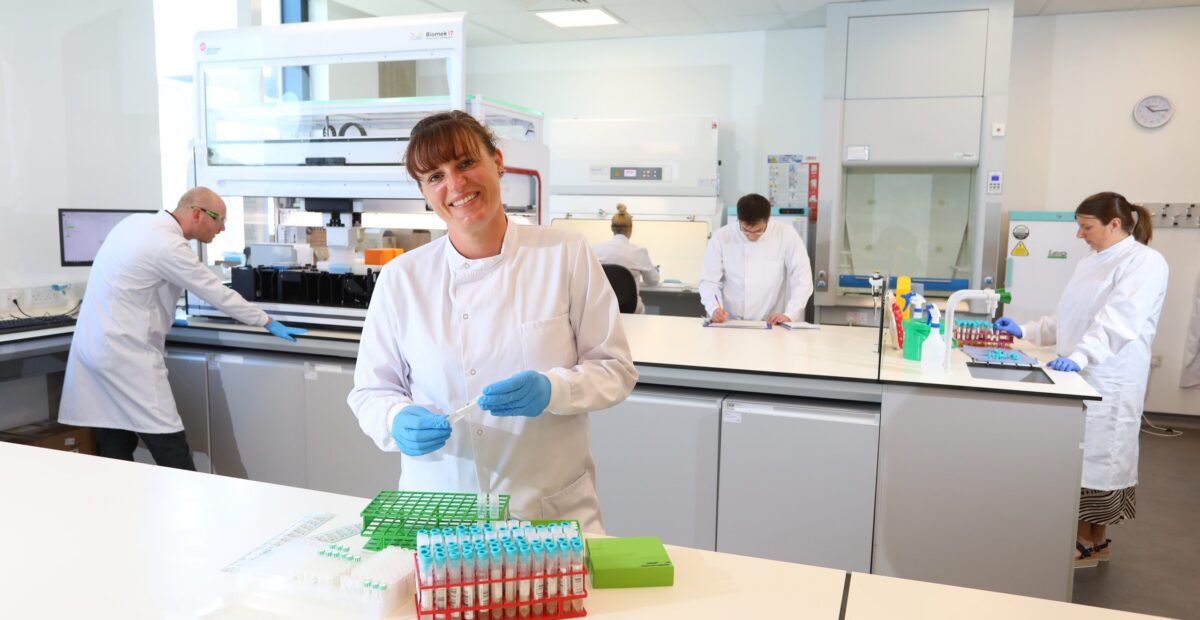 The inside of the innovation lab with scientists at work, and a female scientist smiling at the front with some test tubes.