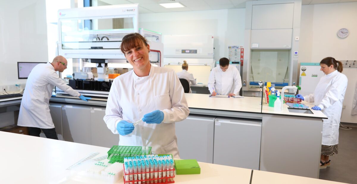 The image shows the inside of the innnovation lab with scientists at work, and a female scientist smiling at the front with some test tubes.