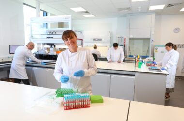 The image shows the inside of the innnovation lab with scientists at work, and a female scientist smiling at the front with some test tubes.