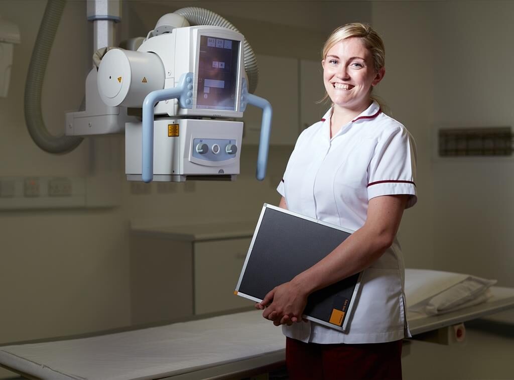 A radiographer wearing uniform and holding a black board, standing next to the X-ray machine.