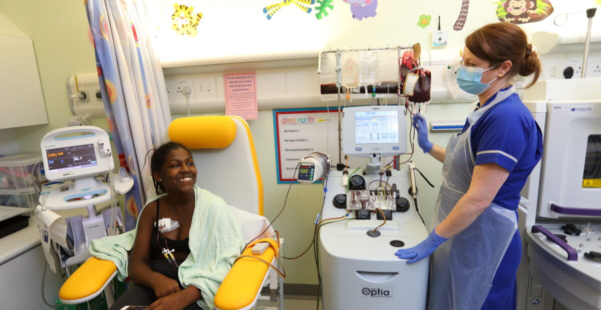 A lady sits in a chair on a ward with a nurse and receives an infusion. The nurse is wearing her blue uniform as well as a face mask and clear apron.
