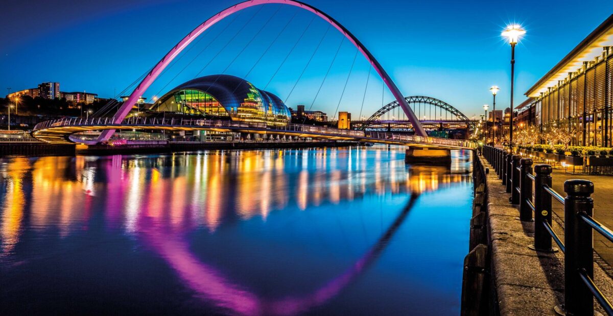 A view of Newcastle Quayside in the ;ate evening with the millennium bridge lit up pink with the sun setting in the distance.