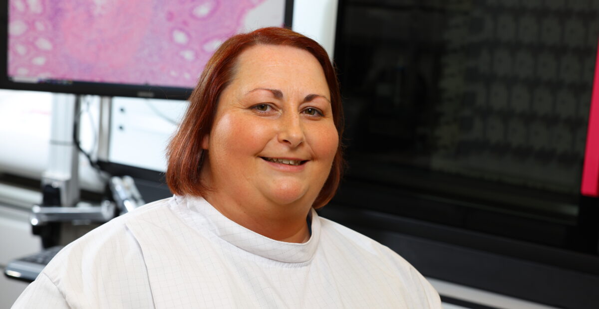 A scientist smiling sitting in front of a screen with a microscopic image displayed.