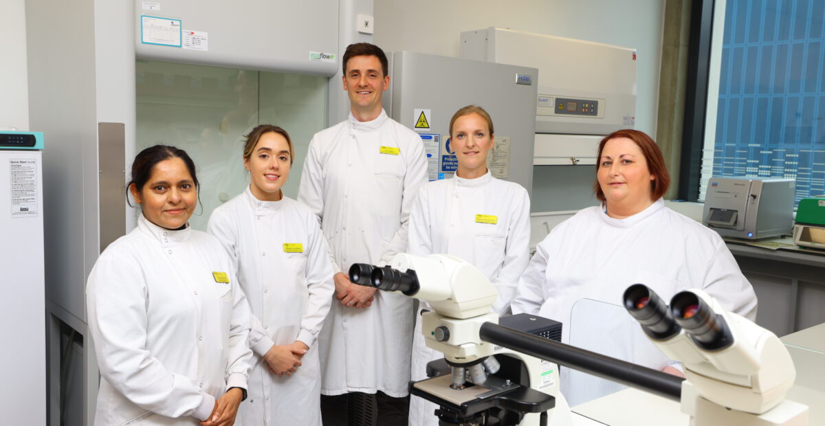 Five scientists at The Biosphere wearing white lab coats and yellow identification badges. They are smiling for the photo.
