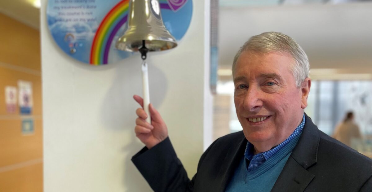 A man ringing the bell on a cancer ward. There is a rainbow on the wall next to the bell.