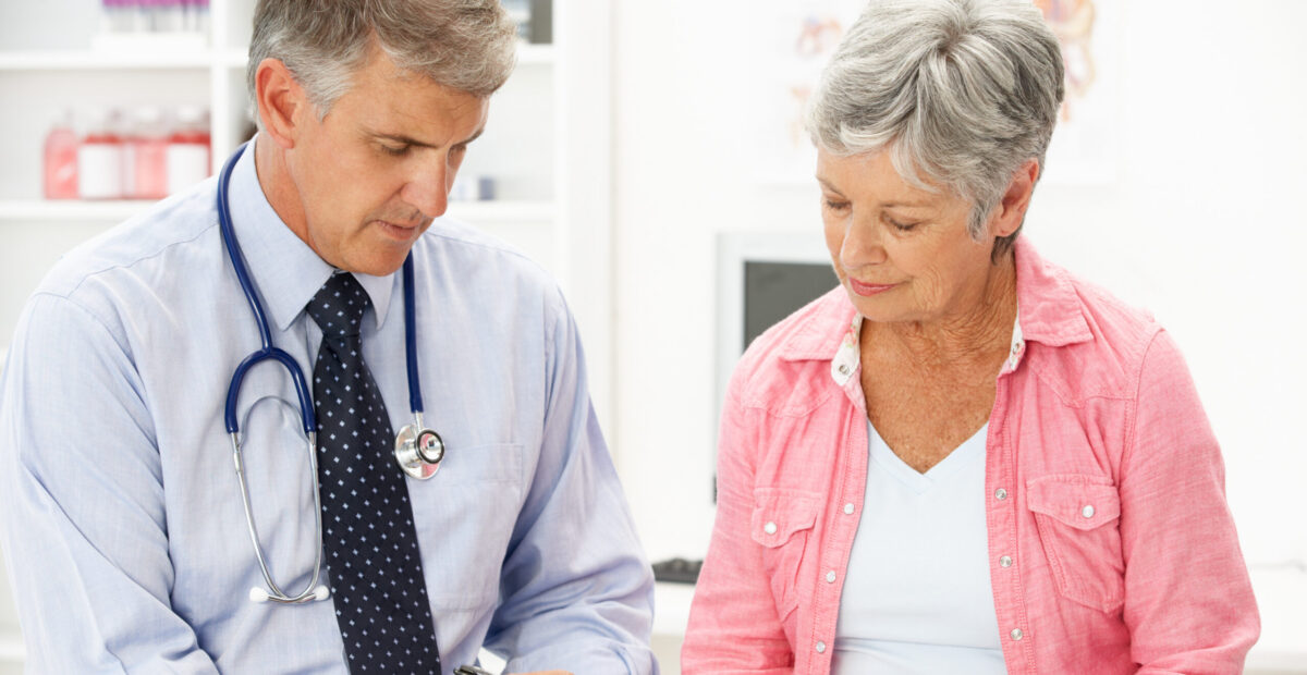 A doctor with a stethoscope consulting with his patient. There is a poster with a diagram of the human body on the wall in the background.