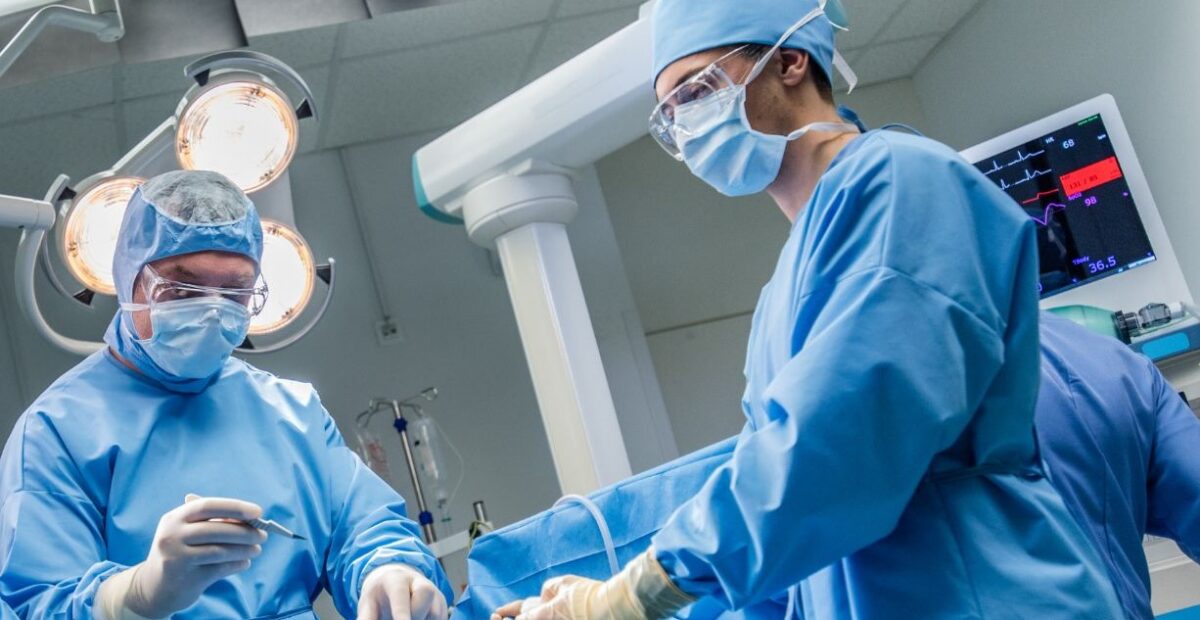 Surgeons operating in theatre wearing blue scrubs and PPE. There is a large surgical light behind one of the surgeons.