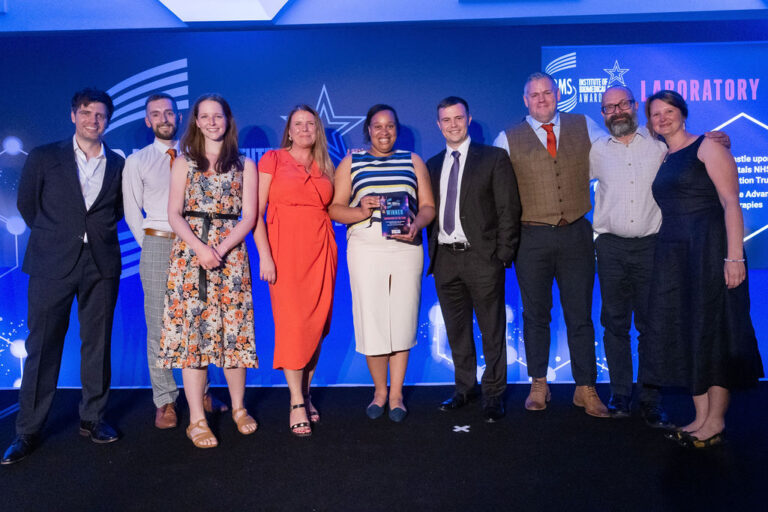 A group of people at the IBMS awards with an award, standing in front of a blue background with a logo on.