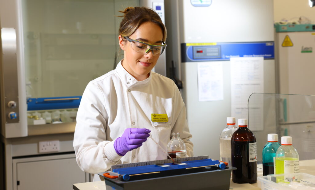 A scientist doing a test wearing a white lab coat and a yellow identification badge.