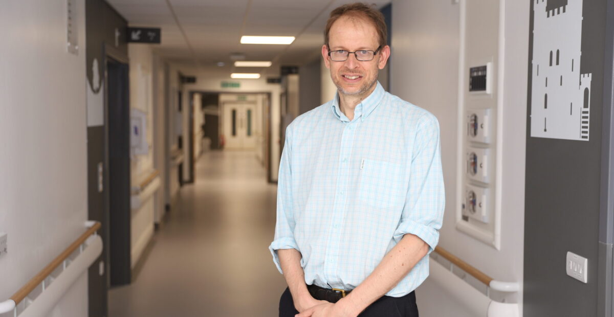 A man in a hospital corridor wearing a baby blue shirt and glasses.