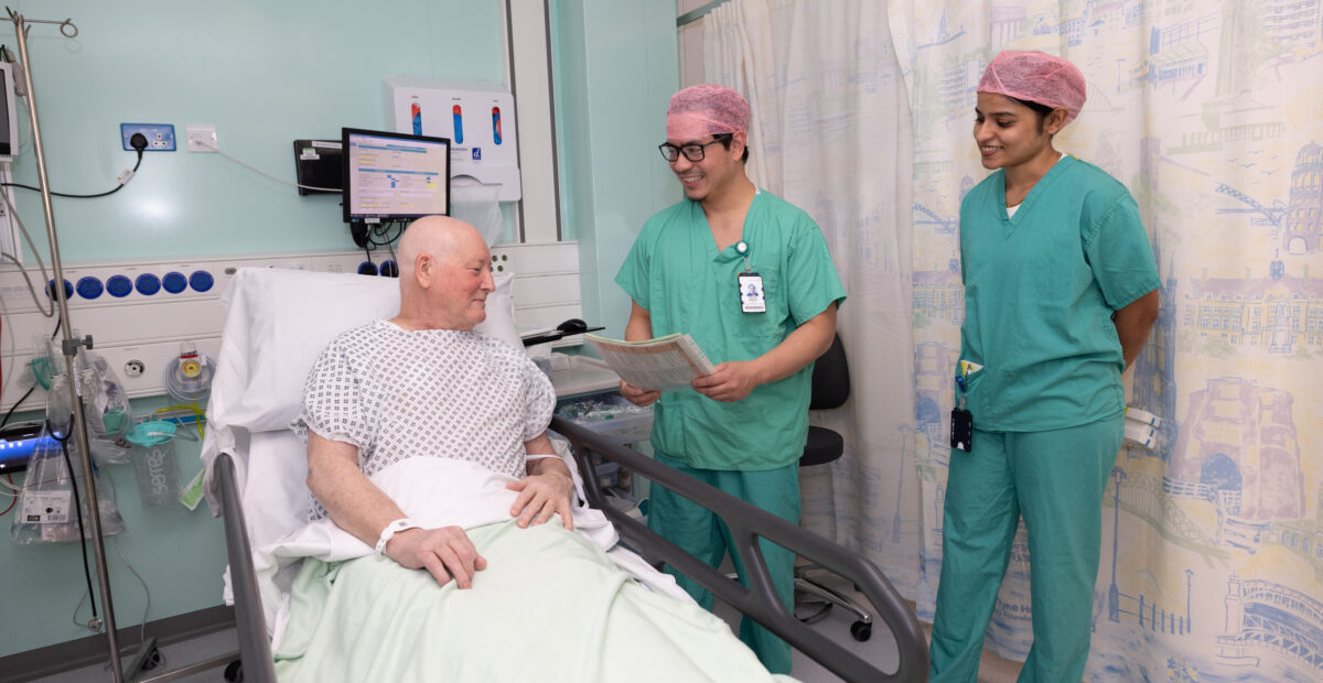 Two doctors, wearing uniforms and protective hats, standing next to a patient in bed, who is in a hospital gown.