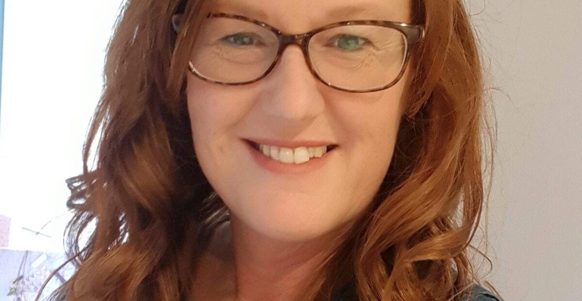 A woman with red hair and glasses smiling.