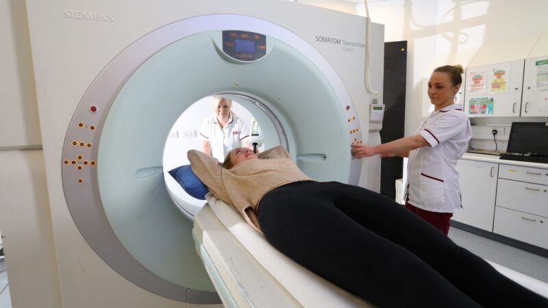 Two staff members wearing white uniform assisting a patient into a CT scan.