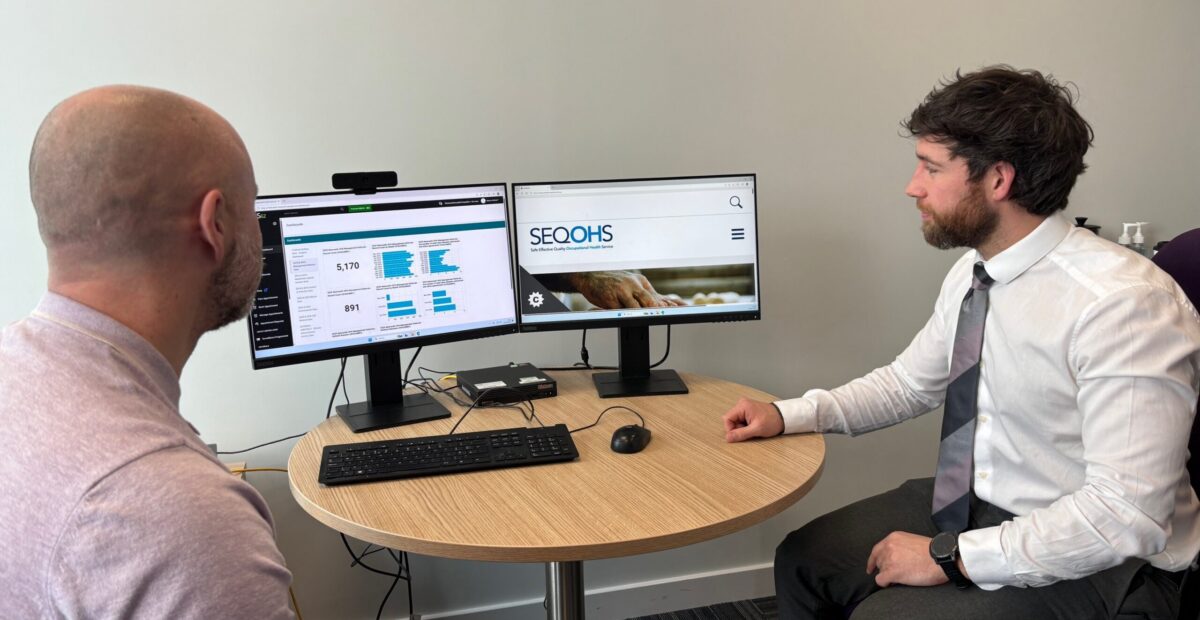 Two men sat at a desk discussing information which is displayed on a computer screen.