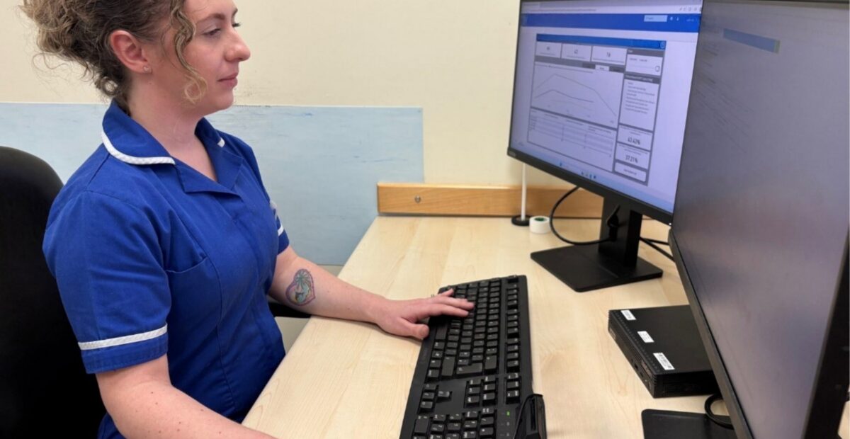 A nurse in uniform sits at a desk in front of a computer screen showing the software