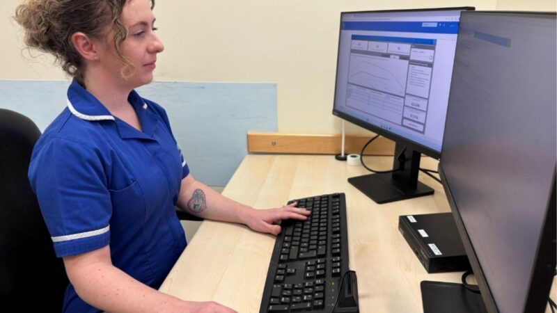 A nurse in uniform sits at a desk in front of a computer screen showing the software