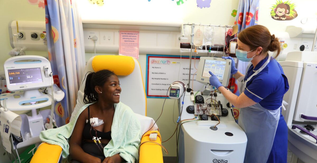 A nurse talking to a patient whilst administering a blood transfusion. The nurse is wearing a blue uniform and a protective apron and face mask. The patient is smiling.
