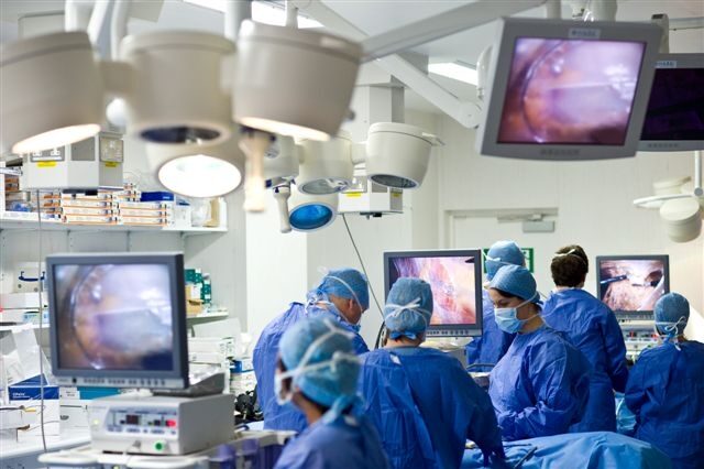 A team of surgeons carrying out an operation. There is an image displayed on multiple screens in the operating room which showing that is being seen.