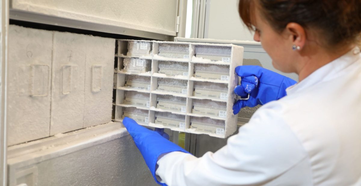 The image shows a female scientist in a white coat and protective gloves taking out samples from the biobank.