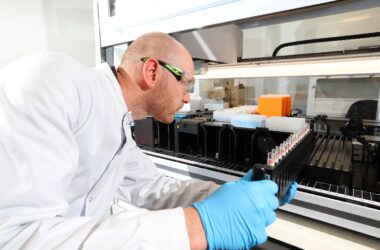 The image shows a male scientist putting a rack of test tubes into a large piece of lab equipment.