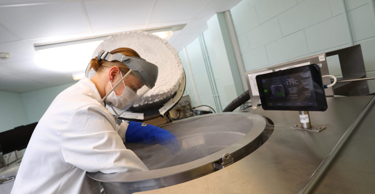A scientist in protective gear leans over a vat with the lid open