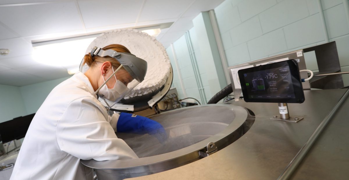 The image shows a female scientist in protective mask and lab coat leaning over a tank with the lid open.
